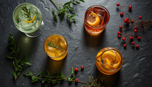 A top-down view of four iced cocktails in glasses with yaupon holly plant leaves, lemon, and red berries on dark slate.