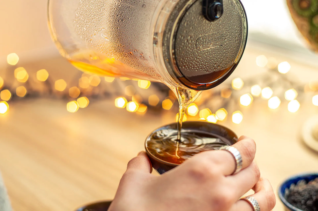 A glass teapot pouring hot, dark amber tea into a small ceramic cup, showing the initial preparation step for making yaupon iced tea with warm bokeh lights in the background.