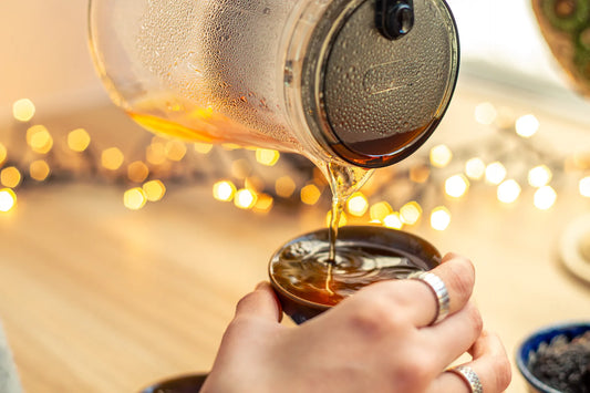 A glass teapot pouring hot, dark amber tea into a small ceramic cup, showing the initial preparation step for making yaupon iced tea with warm bokeh lights in the background.