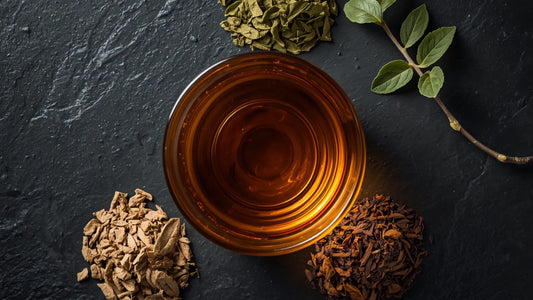 A top-down view of a clear glass cup filled with brewed tea surrounded by piles of dried green, light roasted, and dark roasted loose leaves on a dark slate background, analyzing Catawba yaupon vs other yaupon tea brands for higher antioxidant levels.