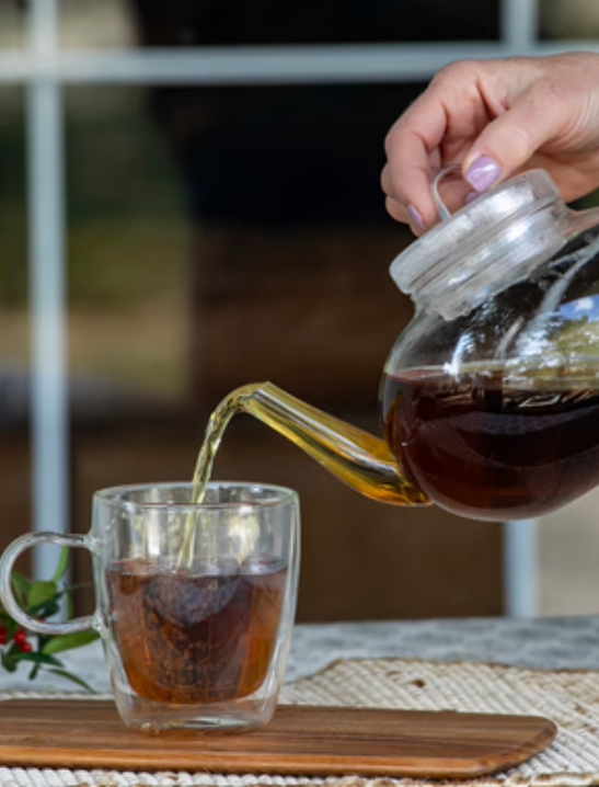 A hand pouring freshly brewed dark yaupon tea from a glass teapot into a clear glass mug on a wooden coaster.
