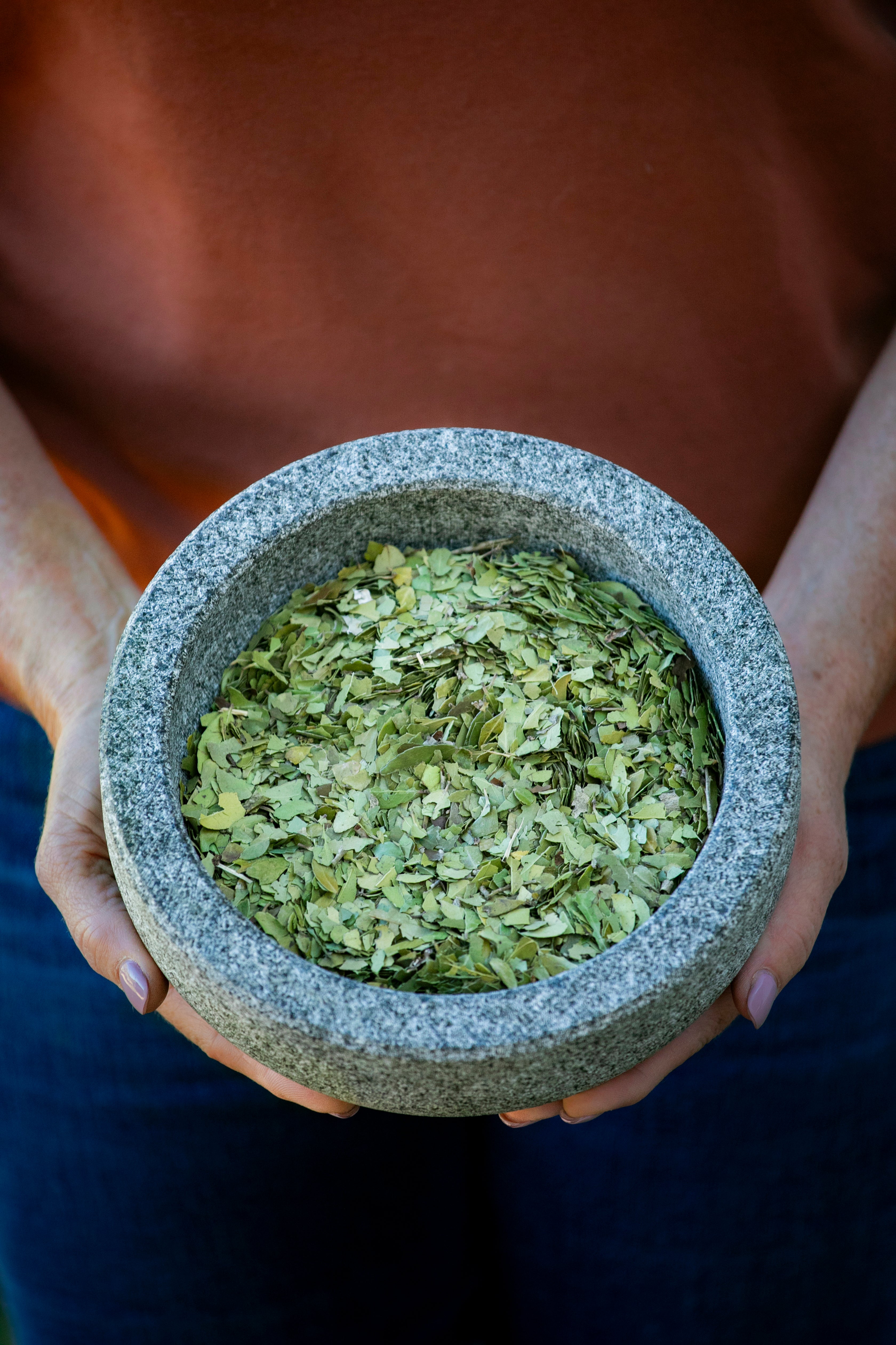 A person holding a stone mortar filled with crushed green herbal tea leaves, representing natural ingredients and craftsmanship.