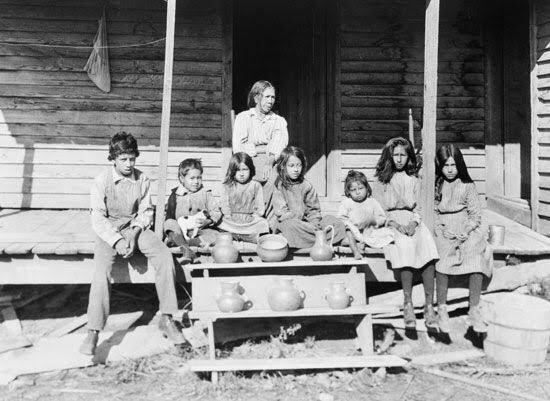 A historical black-and-white photograph of Catawba women and children sitting on a porch with their traditional hand-built pottery displayed on steps in the foreground.