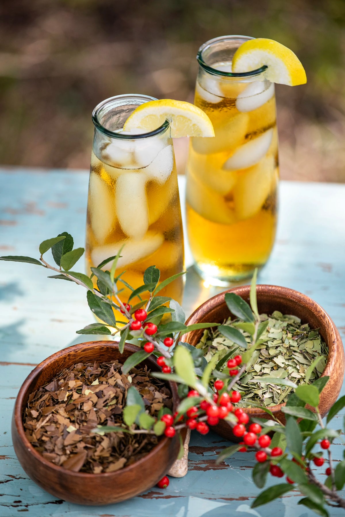 Two tall glasses of iced Yaupon tea garnished with lemon slices, positioned behind two rustic wooden bowls containing different types of loose-leaf Yaupon tea and a sprig of fresh Yaupon holly with bright red berries.