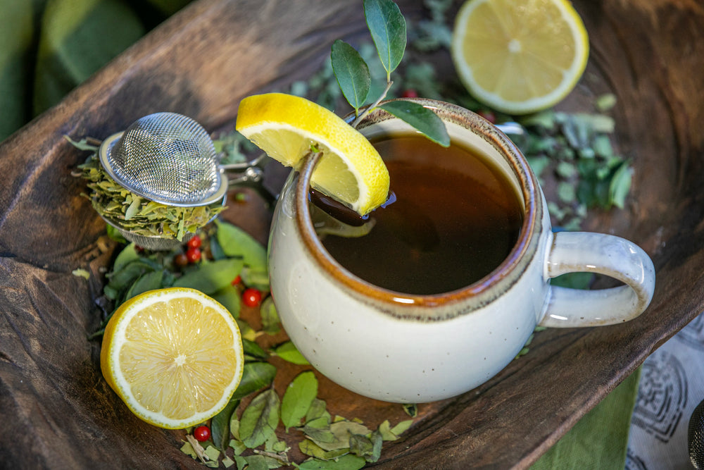 A ceramic mug of dark tea garnished with a lemon slice, nestled in a wooden bowl filled with loose-leaf Yaupon holly, fresh lemon halves, and a metal tea infuser.