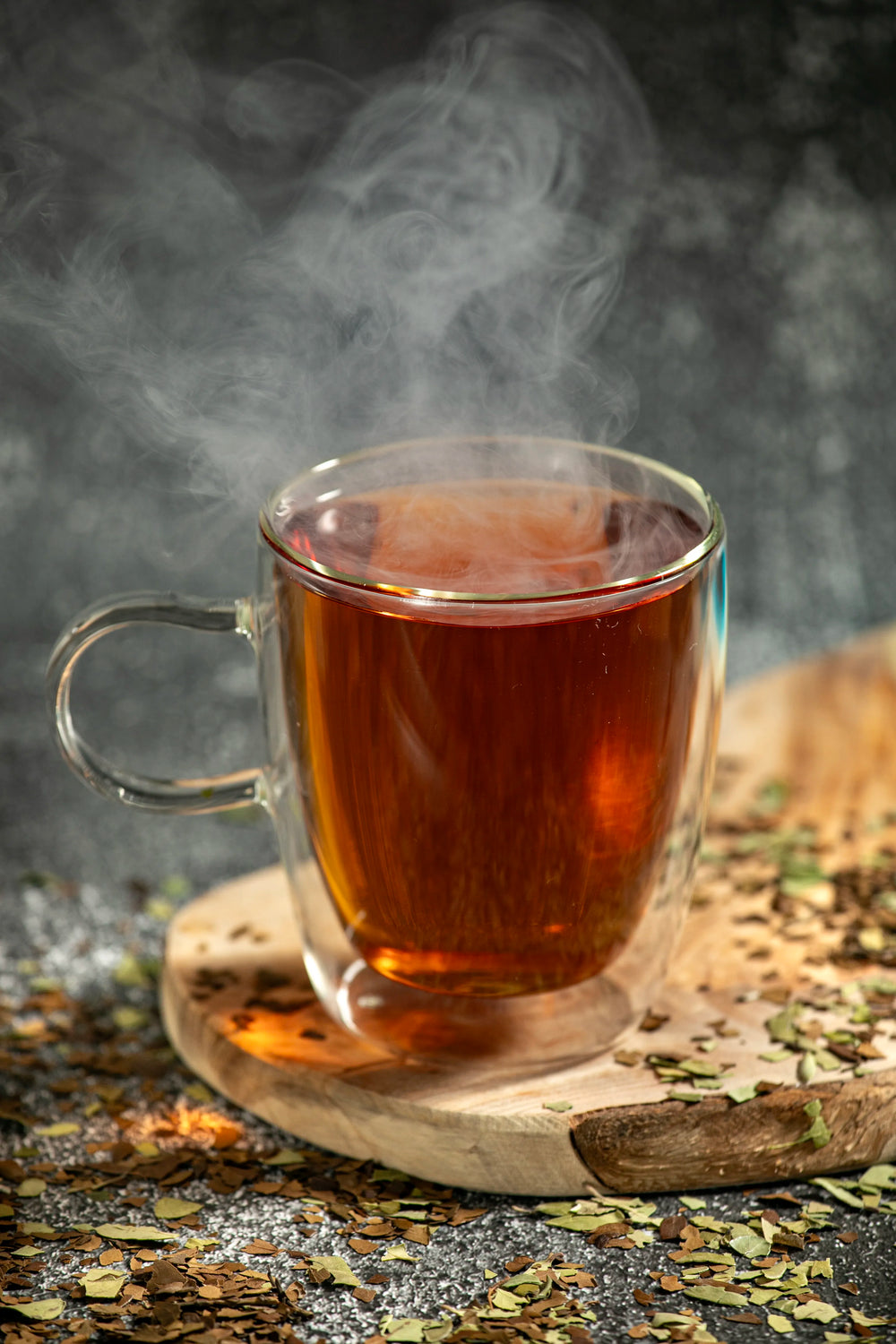 A steaming, double-walled glass mug containing freshly brewed, dark amber Yaupon tea—North America's only native caffeinated plant—placed on a rustic wooden board scattered with loose, dried leaves.