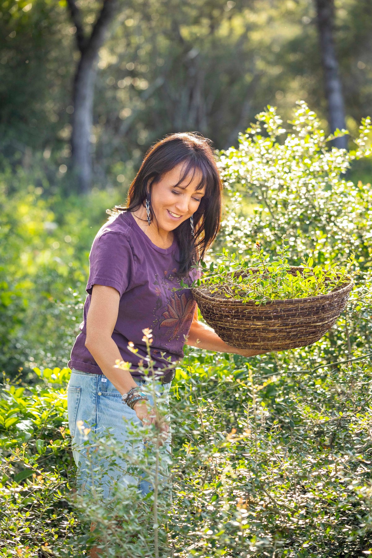 A woman wearing a purple t-shirt and jeans smiles while gathering fresh Yaupon holly sprigs into a traditional woven basket in a sun-drenched, natural field.