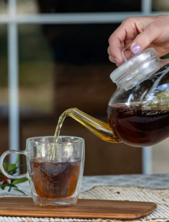 A hand pouring steaming, dark amber Yaupon tea from a clear glass teapot into a double-walled glass mug, highlighting the warm, inviting preparation of North America's only native caffeinated plant.